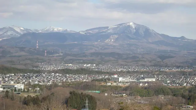 The image depicts a mountainous landscape with snow-capped peaks in the background. In the foreground, there appears to be a densely populated area, likely a town or city, with various buildings and infrastructure visible. The landscape is lush with trees, and there are power lines stretching across the view. The overall scene suggests a mix of urban and natural environments, potentially in a region with a temperate climate.

Image Credits: Wikimedia / User: Wkatsuhiro / Public domain