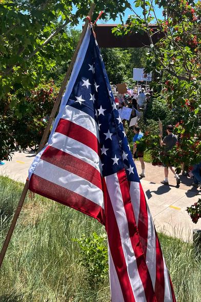 A US flag draped by the side of a path in a park, with a peek of a large gathering of people visible through a break in the background trees, all under bright, sunny skies.