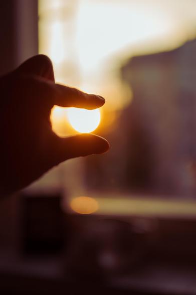 A hand silhouetted against a bright sunset sky, fingers positioned as if pinching the sun, creating a visual effect of holding the sun between thumb and forefinger. The background is softly blurred with warm golden tones.
