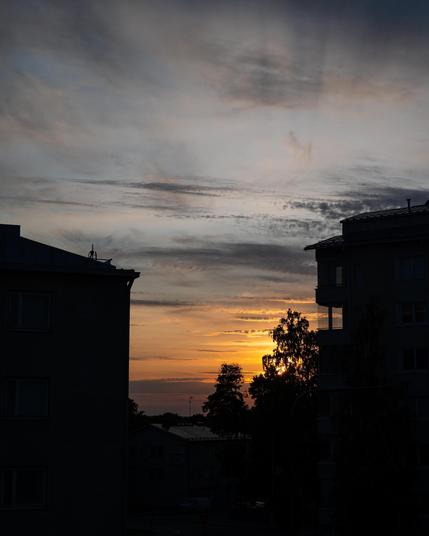 Sunset with an orange and yellow sky, partially obscured by wispy clouds. Silhouetted buildings frame the scene on either side, and tree outlines are visible in the foreground.