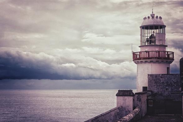 A dramatic, wide shot of a white and red lighthouse standing on a cliff edge, overlooking a dark, choppy sea under a foreboding, cloudy sky. The clouds in the distance are heavy and dark, suggesting a storm. The overall tone of the image is melancholic and powerful, with a hint of purple in the sky and water.