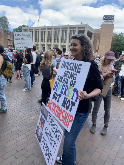 Professor holds sign reading “imagine being afraid of diversity but not dictatorship” - at her feet is another reading “Impeach, convict, remove”