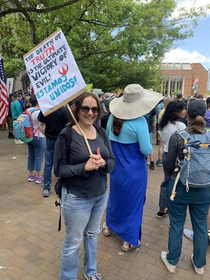 Latina holds sign reading “The death of truth is the ultimate victory of evil” with the Andor symbol next to it. Below it says “Estamos Unidos!” - à large American flag is seen in the background