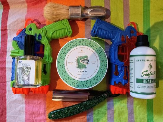 (Le Chat Mistral description)

The image features a collection of grooming and personal care items arranged on a striped background. At the centre, there is a round tin of shave soap with a dinosaur illustration, labelled "RAWR." To the left, there is a bottle of Eau de Toilette with a hippopotamus illustration, and a green toy water gun. To the right, there is a bottle of Seaforth! Heather aftershave. Below these items, there is a straight razor with a glittery handle and a shaving brush with a wooden handle. The items are colourful and varied, creating a vibrant display.