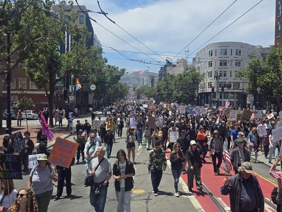 A view up Market Street San Francisco looking towards Twin Peaks. The street is filled with people holding signs to protest the Trump administration. The street is filled with people as far away as the eye can see.