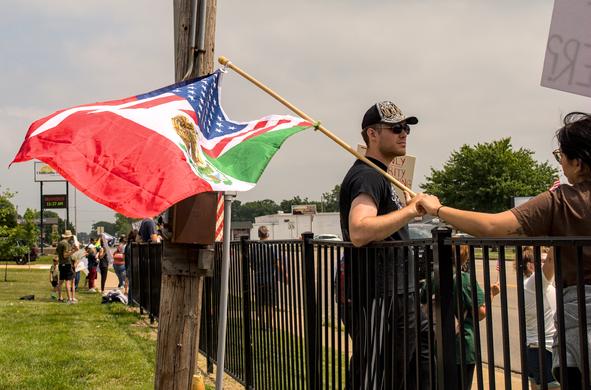 Man standing against a fence waving an American and Mexican flag. He is wearing a hat and sunglasses and holding the hand of a woman next to him.