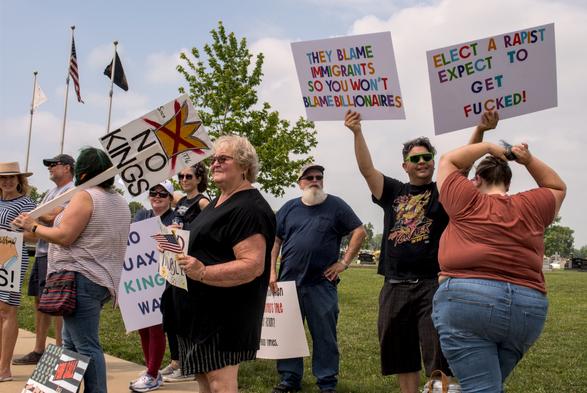 Various protestors holding signs. A woman has a sign resting on her shoulder that reads "No kings" with a picture of a crossed out crown. Another man is holding 2 signs. One reads "They blame immigrants so you won't blame billionaires" and the other reads "Elect a rapist expect to get fucked!"