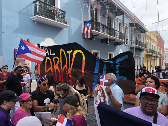 Crowd of people in front of balconied buildings of San Juan. The crowd waves Puerto Rican and Dominican flags. Large banner sits in front of the fortaleza which reads “Pal Carajo ICE”