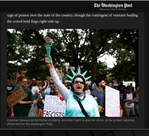 man sieht eine Frau mit Sonnenbrille auf einer no-kigs-demonstration, die sich erkennbar mit Srahlenkranz und Fackel als "Statue of Liberty" zeigt und das in einem weiß-blauen Kleid. dahinter kann man auf einem  Demoplakat lesen: "Who loves Military Parades? Facists"