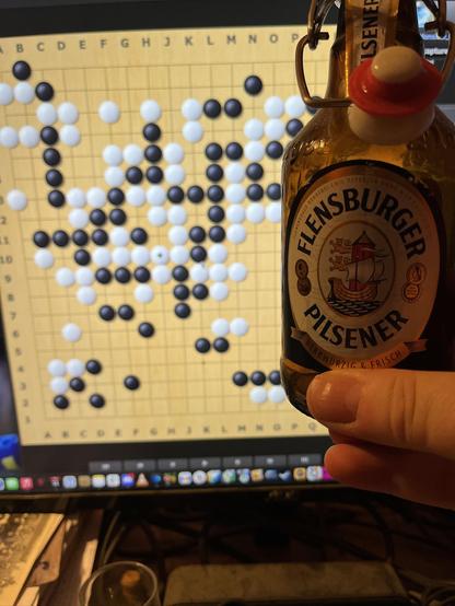 I am holding up an empty bottle of Flensburger Pils in front of a computer monitor, on which I am playing Go. I'm white, and have captured a humungous black group. Hurrah for me!