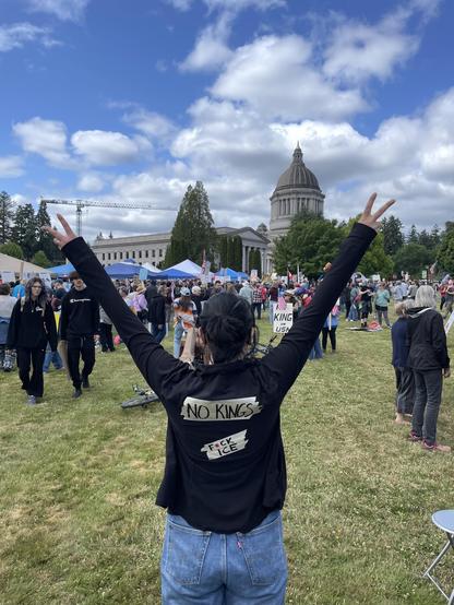 A person standing in front of the Washington State Capitol, arms raised in peace signs with “No Kings” and “F*ck ICE” written on their back.