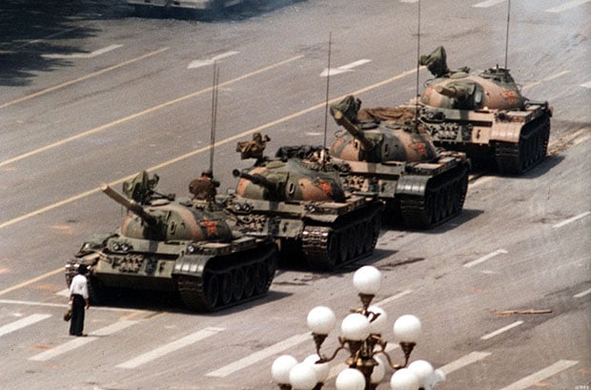 A lone man in a white shirt and black pants carrying two shopping bags stands in front of a column of military tanks blocking their path near Tiananmen Square Beijing June 5 1989