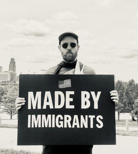 A bearded person (me) with sunglasses and a hat is holding a large sign that says made by immigrants with a USA flag