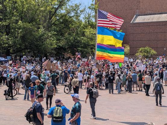 A photo of a man holding a large pole with three flags on it in fronts of a crowd. From top to bottom the flags are US, Ukraine, and Pride