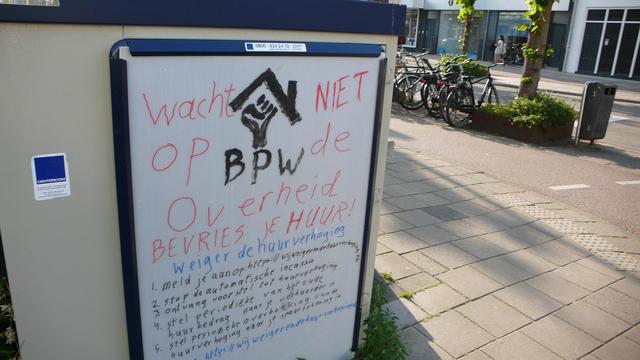 Wit bord op beige kast met straat en geparkeerde fietsen en bomen, prullenbak en egbouw aan de overkant.
Wit bord met zwart een vuist onder een woondak BPX
In rood: Wacht niet op de overheid. bevries je huur!
In blauw: weiger de huurverhoging
in zwart: 
1. Meld je aan!
2. Stop de automatische huurincasso
3. Ontvangst voorstel tot huurverhoging
4. Stel een periodieke overboeking in van het oude huurbedrag naar je verhuurder
5. Stel een periodieke overboeking in van de huurverhoging naar je spaarrekening. 
In blauw de website