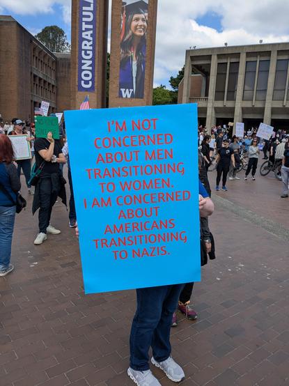 A person at a protest holding a sign that reads "I'm not concerned about men transitioning to women. I'm concerned about Americans transitioning to Nazis."