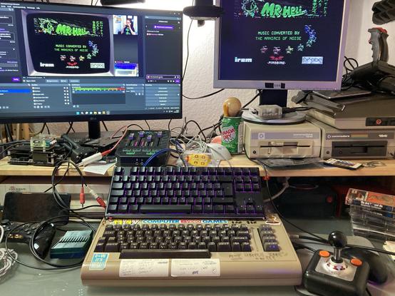My streaming setup with a C64 running the Mr. Heli title screen. There’s two screens connected, the left one shows the OBS streaming screen from my PC and the right one shows the video output from the C64. A Tac-2 joystick is visible in the lower right corner. There’s a lot of cables and debris all around that needs tidied up.