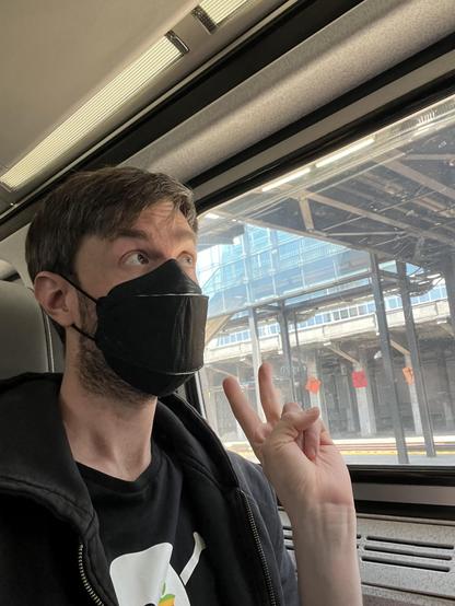 Photo of me in a black mask giving a peace sign and looking out the window of an amfleet car stopped at king st station in seattle