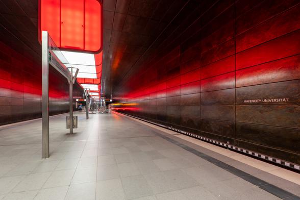 U-Bahnhof HafenCity Universität in Hamburg, Bahnsteighalle mit dem Mittelbahnsteig. Auf dem rechten Gleis verlässt ein Zug der Linie U4 in Richtung Elbbrücken die Station. Die obere Hälfte der Station ist rot beleuchtet.