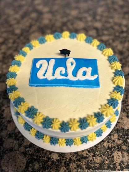 A round cake decorated with a white frosting base. The top features a blue rectangular design with the word "UCLA" in white cursive. Surrounding the top edge is a border of blue and yellow star-shaped icing. A small black graduation cap is stuck on top of the box holding the lettering