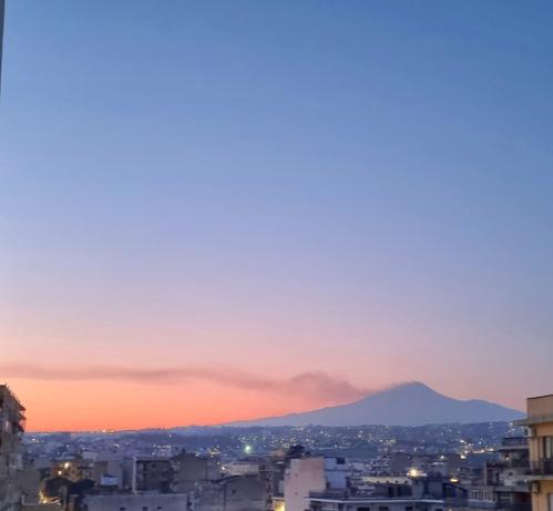 Tra i colori pastello del tramonto, l'Etna fuma, lasciando una lunga scia nel cielo sereno