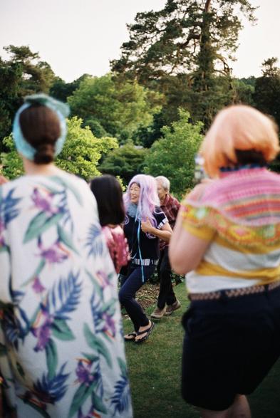 A lively outdoor gathering featuring people wearing colorful outfits and wigs. In the foreground, one individual with long purple hair is smiling and dancing, while others in vibrant clothing are partially visible. The background includes lush greenery and trees.
