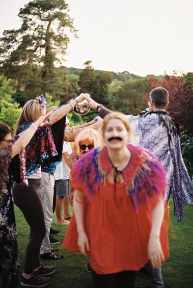 A group of people in colorful costumes and accessories are dancing in a garden setting. Some are wearing capes and others have novelty glasses or wigs. The atmosphere appears festive and joyful, with greenery in the background and a warm sunset light.