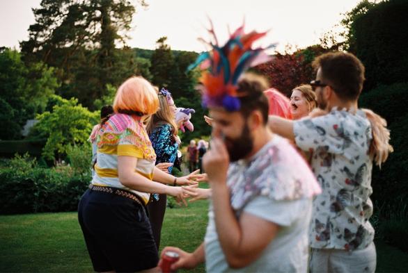 A lively outdoor scene featuring a group of people dancing and celebrating. Attendees are dressed in colorful and playful outfits, including wigs and vibrant accessories, surrounded by greenery. The atmosphere is joyful and festive.
