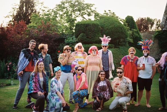 A colorful group of people dressed in eclectic costumes and accessories, posing playfully outdoors. The scene features various hats, wigs, and oversized sunglasses, alongside items like a plush toy and a chicken prop. The background includes lush greenery and flowers.
