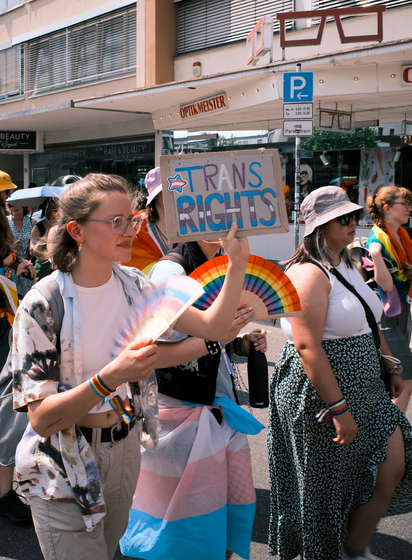 Personen mit Fächern in trans und regenbogen Farben halten ein Schild hoch mit dem Text: Trans Rights