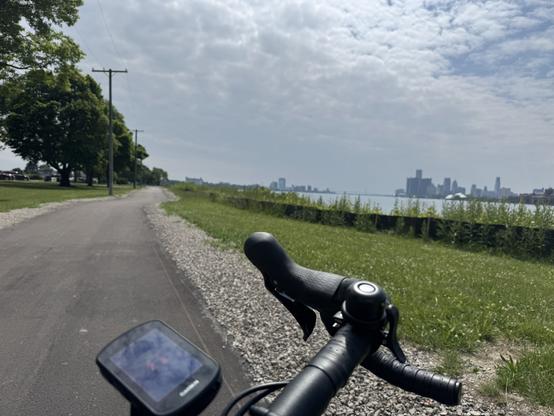 A view over the handlebars of a road bike. The Detroit skyline can be seen in the distance under a blue sky with light, wispy clouds.