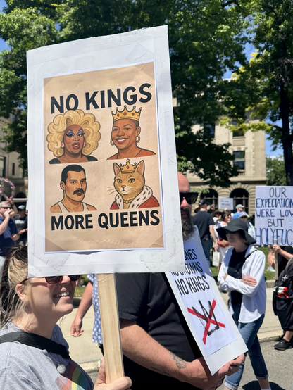 Sign reading , No Kings More Queens with four pictures of “queens, including Freddie Mercury. In back ground is sign, Our expectations were low but holy fuck