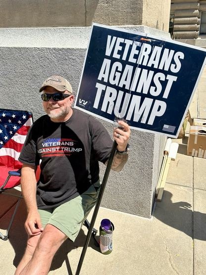 A smiling white man wearing a tshirt, Veterans Against Trump and holding a sign saying the same