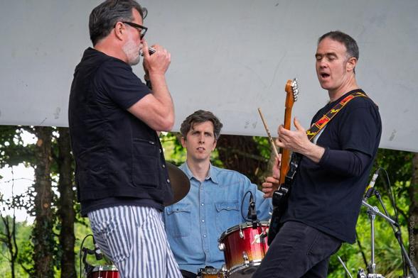 A lively outdoor music performance at Gallagher Fest 2025 in Fitzgerald Park, Cork, featuring a harmonica player in striped trousers, a drummer in a denim shirt, and a guitarist with a colourful strap, all energetically engaged on stage with musical instruments and microphones visible, set against a backdrop of greenery and a white canopy.