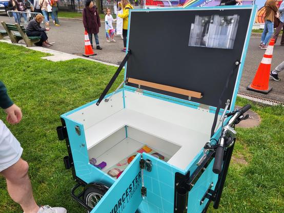 Photo of a cargo bike with livery for the Public Library of Worcester, MA taken on the Boston Common

The lid is raised and a door is swung open, revealing a spacious interior for books and other stuff. The underside of the lid has some holders for pamphlets and the entire thing is a blackboard surface.