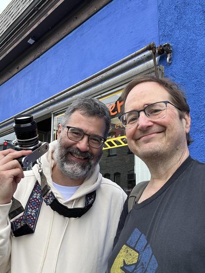 Two middle aged men smile for the camera. One holds up an old film camera, a Nikon FE. He is wearing a white hoodie and has a partially gray beard. The other is wearing a black t-shirt with a blue and yellow fist, which symbolizes solidarity with Ukraine.