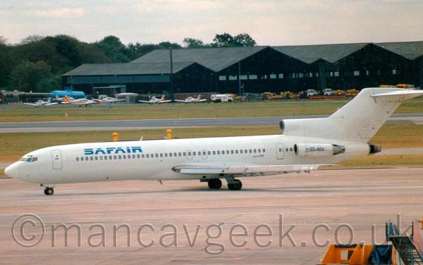 Side view of a 3 engined jet airliner taxiing from right to left.
The plane is  mostly white, with blue "SafAir" titles on the upper forward fuselage, and the registration "ZS-NOV" on the bare metal engine pods mounted on the sides of the rear fuselage.
Grey concrete apron fills the foreground, with grass areas lining the side of a black runway in the background, with black hangars in the distance with white light aircraft and white and yellow vehicles in front.
Cloudy sky with hints of blue fill the rest of the frame.