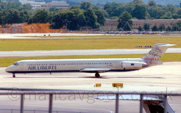 Side view of a twin engined jet airliner with the engines mounted on the sides of the rear fuselage, taxiing from right to left.
The plane is mostly white, with a blue belly, and blue "Air Liberte" titles on the lower forward fuselage.
The tail is mostly white, with blue and red text, mostly French words mostly related to "Liberty".
In the foreground, the top of a railing at the edge of the viewing terraces can be seen at the bottom of the frame, with large areas of grass divided by runways and taxiways in the background, leading up to brown buildings nestled in trees in the distance.