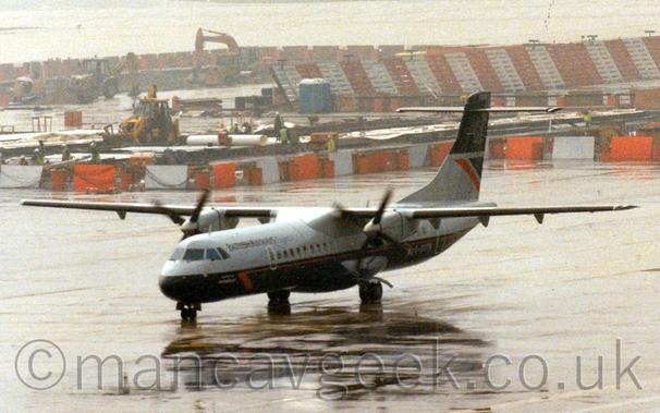 Slightly grainy side view of a high-winged, twin propellor-engined airliner taxiing from right to left.
The plane has grey upper surfaces and a dark blue, almost black belly, with black "British Airways Express" titles on the upper forward fuselage.
Rain-sodden concrete apron fills the foreground, with a rather damp looking building site outlined with white and red striped barriers in the background.