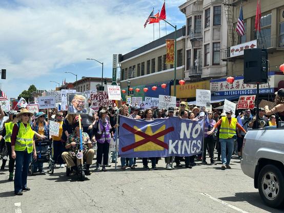 Protest marchers fill a street in Oakland's Chinatown. Many hold signs protesting the Trump administration. The front row of people hold a big banner with a simple picture of a crown with a big red X through it and the words "No Kings"