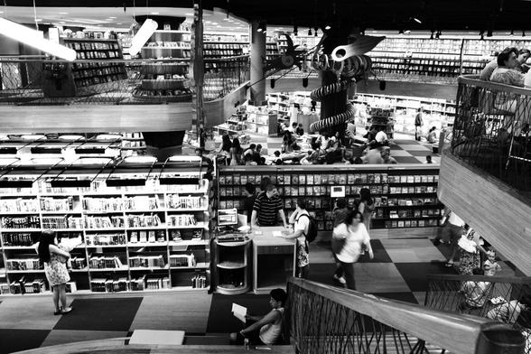A busy bookstore interior featuring multiple levels, with shelves filled with books and DVDs. People are browsing, reading, and interacting at a checkout counter. The image is in black and white, capturing the lively atmosphere of the space.