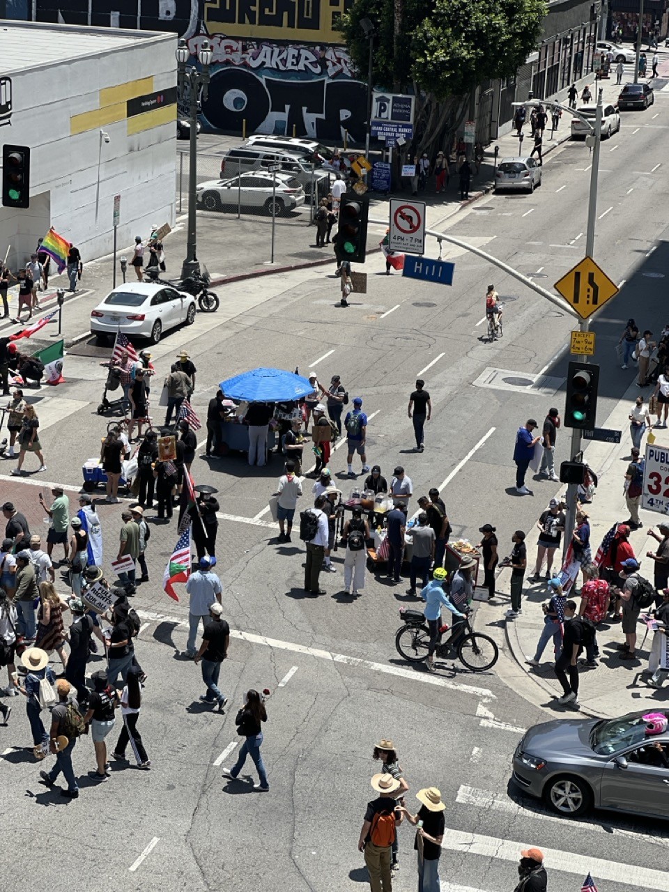 The intersection of Hill and Fourth in downtown Los Angeles during the No Kings march - food vendors are set up in the street selling bacon wrapped hot dogs, aguas frescas, fruit, and other snacks.