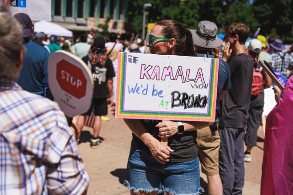 A photo of a woman at a protest holding a sign that reads "If KAMALA won we'd be at BRUNCH" on a sunny afternoon with other protesters visible in the background.