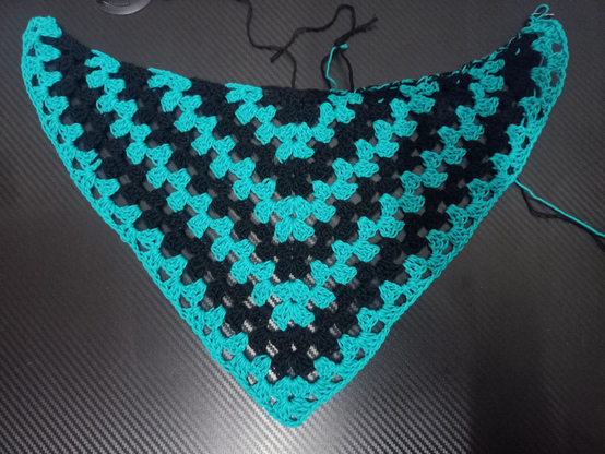 Chal triangular tejido a ganchillo que alterna franjas en forma de V negras y verde azulado, sobre mesa negra.