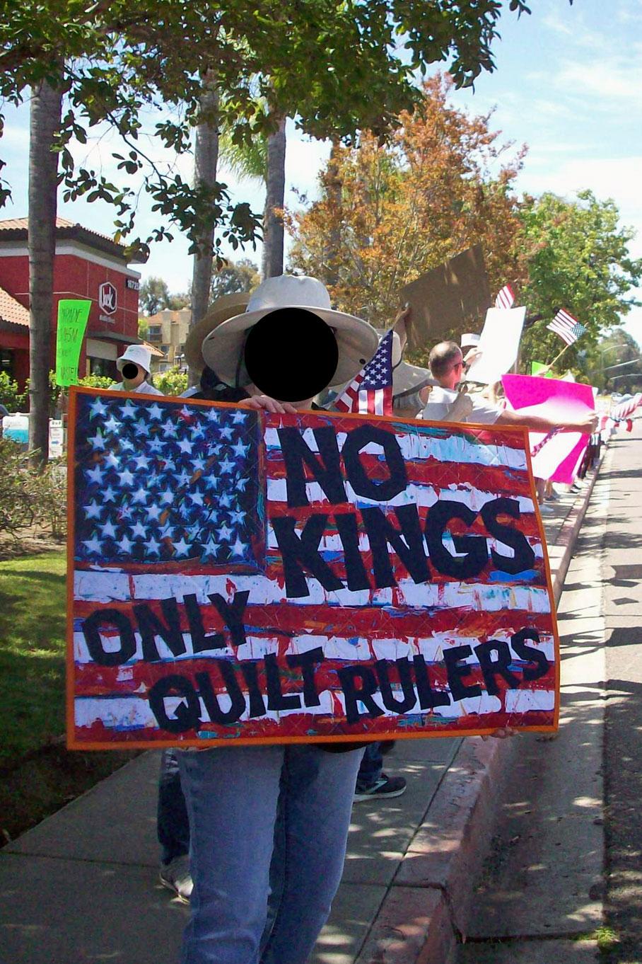 Protestor with a quilted sign that reads No Kings Only Quilt Rulers, the letters are appliquéd on a fabric panel of a colorful, graffitied US flag