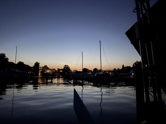Richtung Norden vom Boot aus geblickt. Die Sonne ist untergegangen, aber der Horizont noch orange und nach oben blau. Im Hafen liegen weitere Boote. Unten ist ein Stück vom hochgezogenen Ruderblatt zu sehen. Rechts ist das Solarpaneel Richtung Sonnenaufgang in wenigen Stunden ausgerichtet.