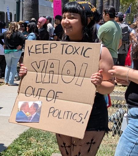 Woman with black hair holds a cardboard sign that says keep toxic you out of politics! At the bottom is a picture of Trump and Musk kissing