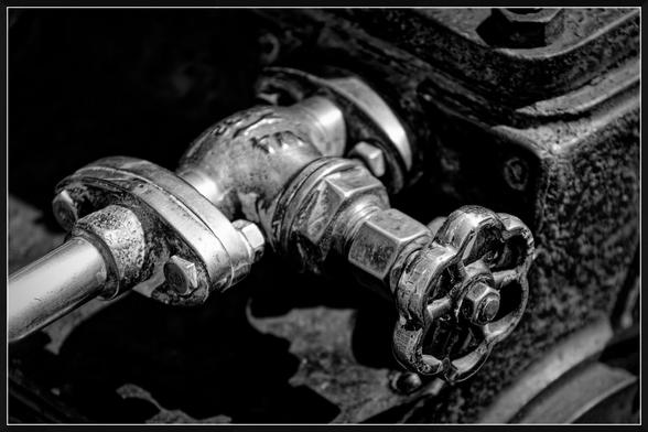 "A close-up view of an industrial metal valve assembly with a wheel-shaped handle, connected to a pipe. The metal surfaces have a textured, aged appearance, showing signs of wear. Bolts and fittings are visible, emphasizing the mechanical structure. The dark background contrasts with the valve, highlighting its intricate details."