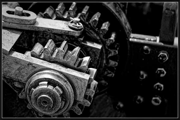 "A close-up black and white photograph of a mechanical gear system. The gears are metallic and show signs of wear and tear, with visible grease and grime. The image captures the intricate details of the gear teeth and the surrounding metal components, including bolts and nuts. The lighting highlights the texture and ruggedness of the machinery, emphasizing the industrial nature of the subject."