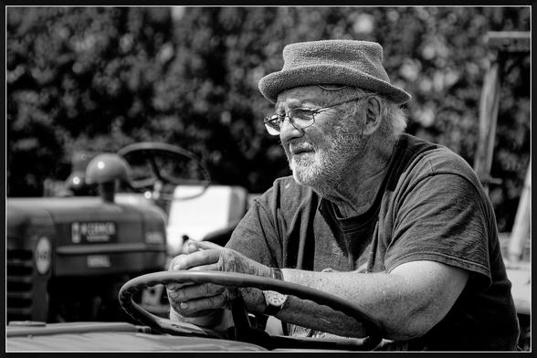 "A black and white photograph of an older individual sitting on a tractor. The person is wearing a hat and a short-sleeved shirt, with hands resting on the steering wheel. In the background, another tractor is visible with the label 'McCormick Farmall' clearly marked. The setting appears outdoors, with trees or foliage framing the scene. The image captures a quiet moment of rest or contemplation, emphasizing the individual's connection to farming or agricultural work."